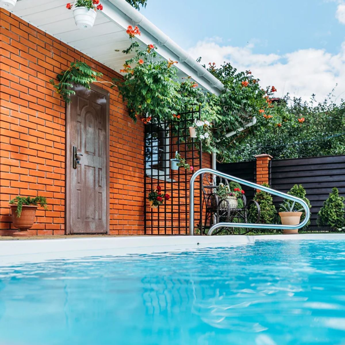 A clean, clear pool next to a bright brick home with lots of hanging blooming plants and shrubs