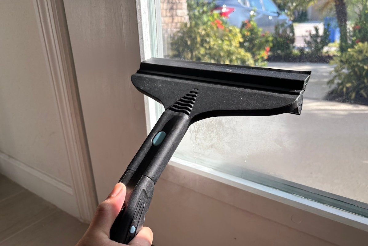 Man uses a squeegee attachment on a steam cleaner to clean a glass window looking out onto a sunny exterior.
