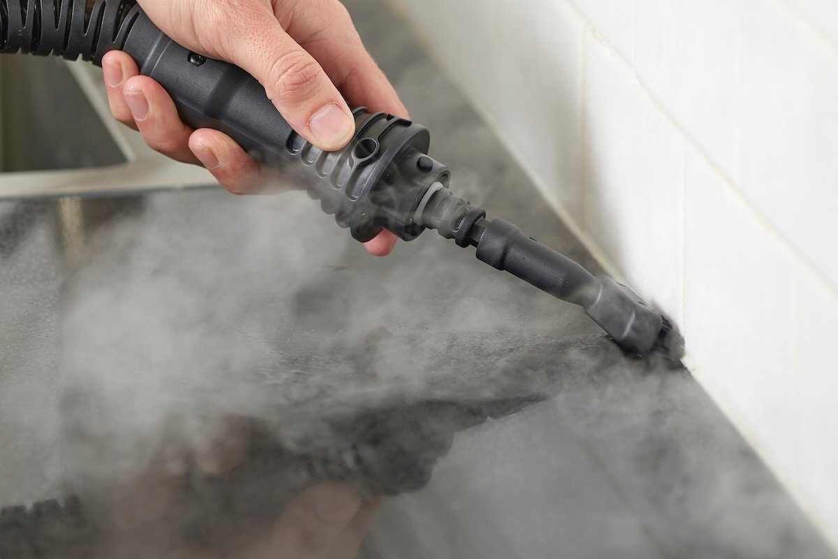 Woman uses a handheld steamer with a narrow attachment to clean grout behind kitchen counter.