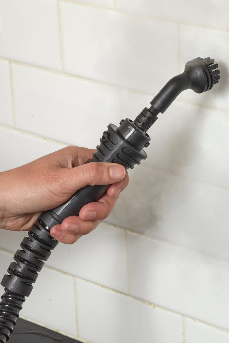 Woman uses a steam cleaner with a wand attachment to clean white subway tiles on a kitchen wall.