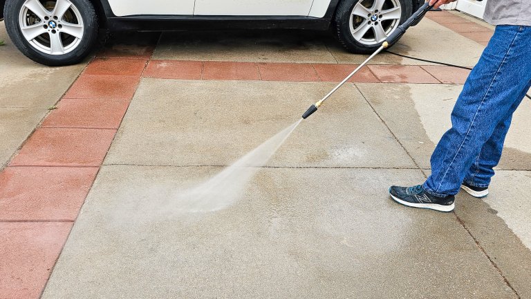 View of man from waist down holding a pressure washer nozzle and pressure washing a driveway, a car in the background.