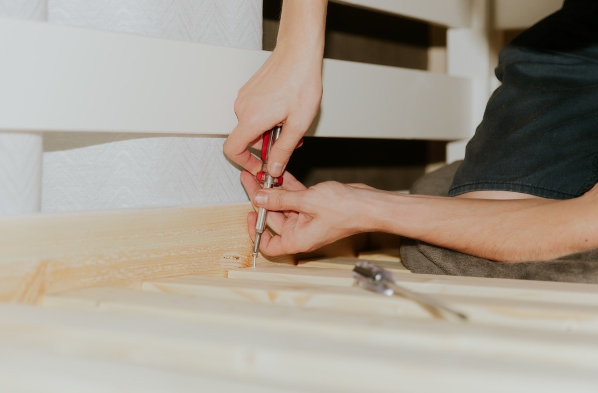 Man sitting sideways on his knees uses a screwdriver to tighten a screw into frame boards on a bunk bed.