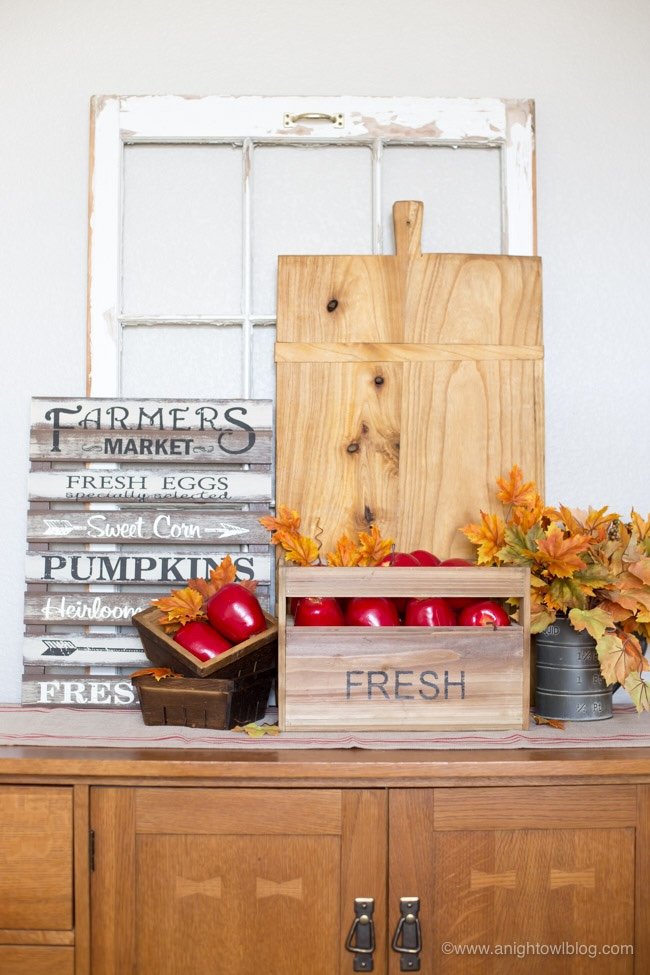 A fall vignette on top of a sideboard, including a painted farmer-like sign, apples in baskets, and a wood board.