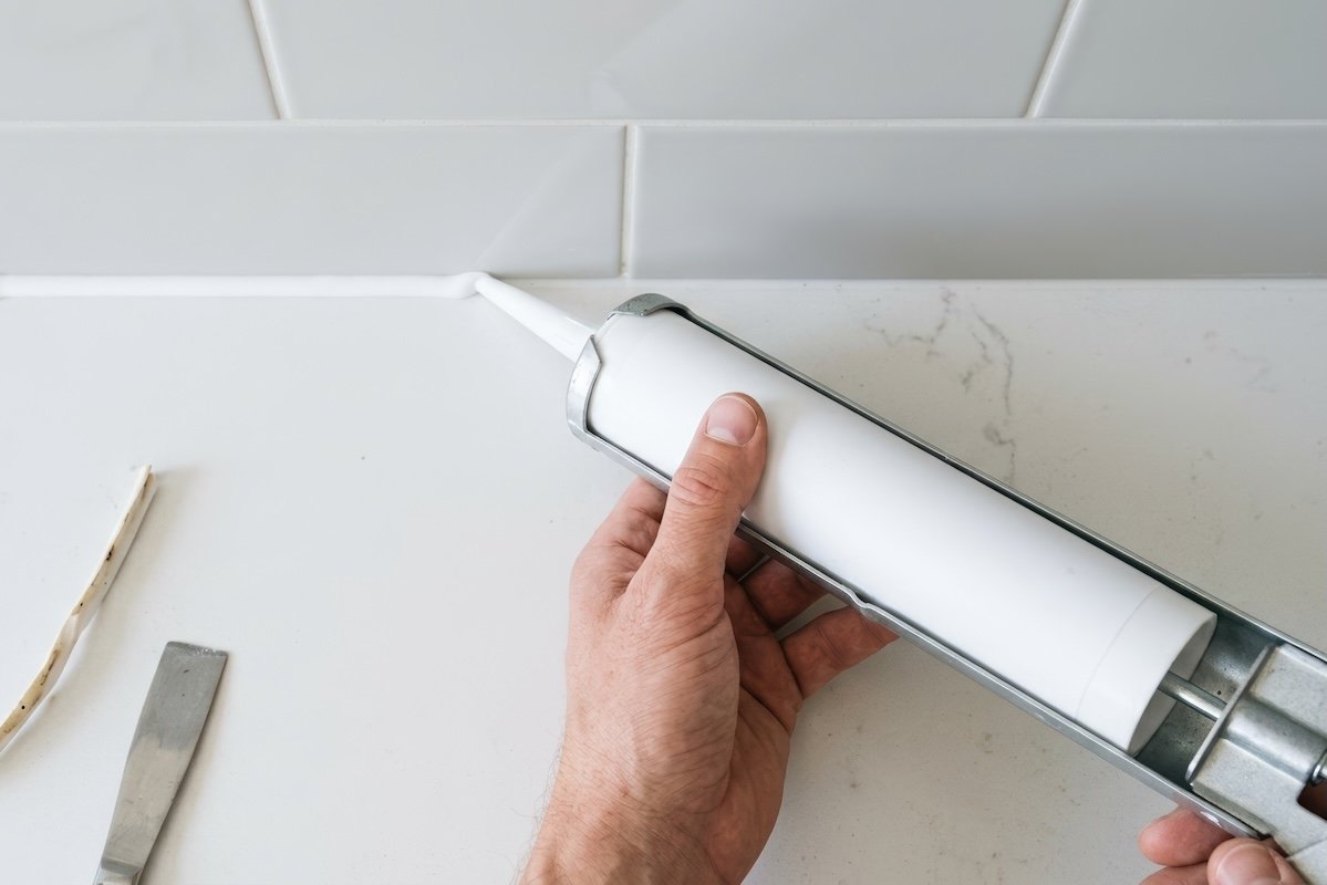 Overhead view of person using caulk to seal seam between white kitchen countertop and light grey backsplash tile.