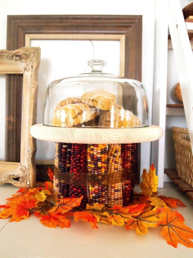 A cake stand made of dried colorful corn cobs holds a wooden plate with pastries under a clear cloche.