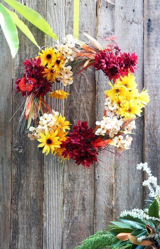 A wicker wreath decorated with autumnal flowers hangs on a wooden fence.
