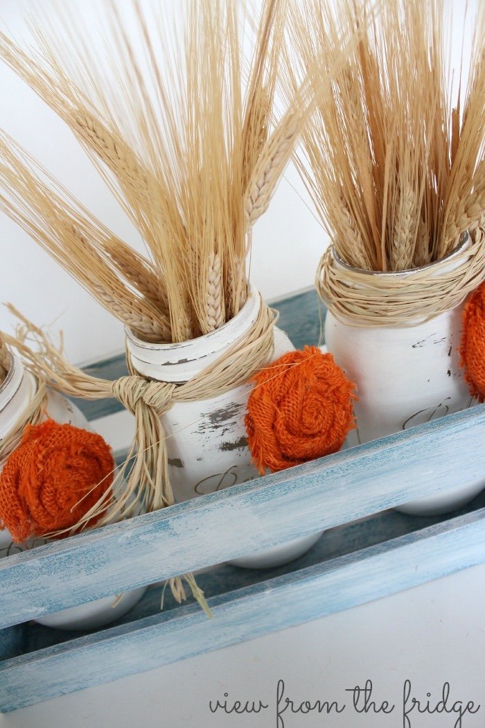 Distressed white Mason jars containing wheat sheafs are decorated with orange pompoms and arranged inside a blue painted wood crate.