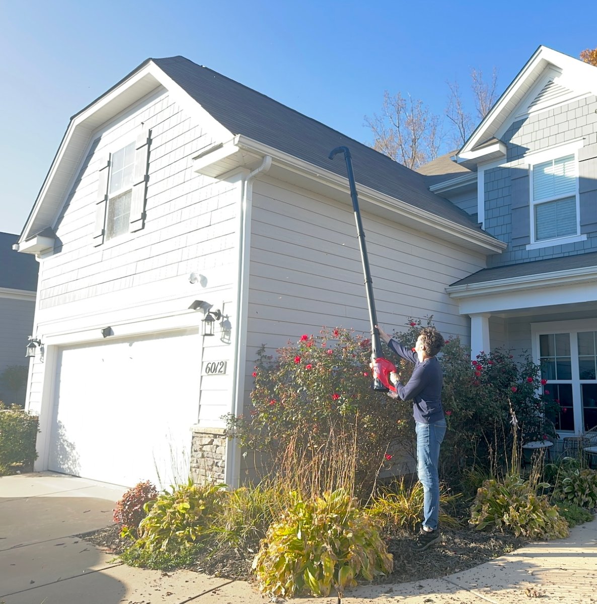 Man uses a gutter cleaning extension for a leaf blower to clean the gutters on a white colonial house.