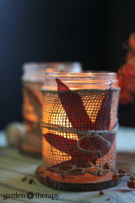 Illuminated votive candle in a glass container decorated with leaves and burlap.