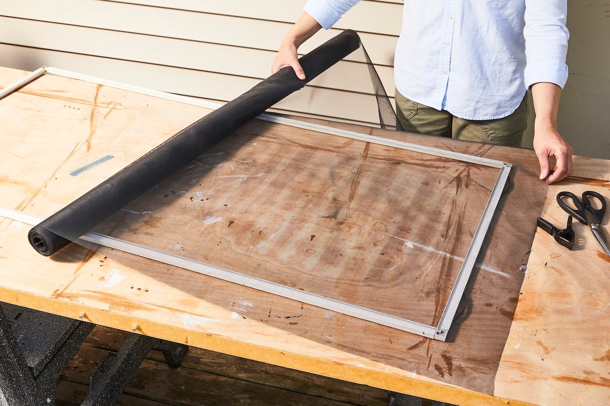 Woman rolls window screen material over a window frame to prepare to cut it.