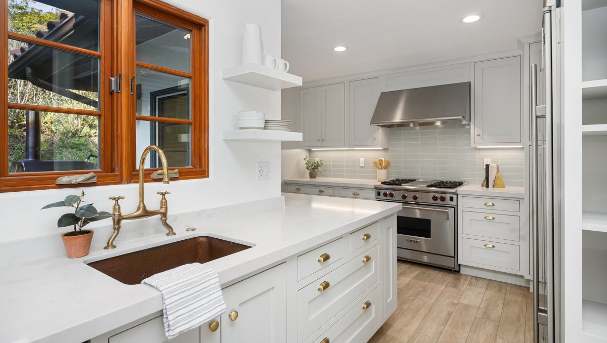 A modern L-shaped white kitchen with a brown window frame behind a brass faucet.