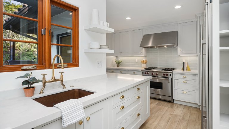 A modern L-shaped white kitchen with a brown window frame behind a brass faucet.