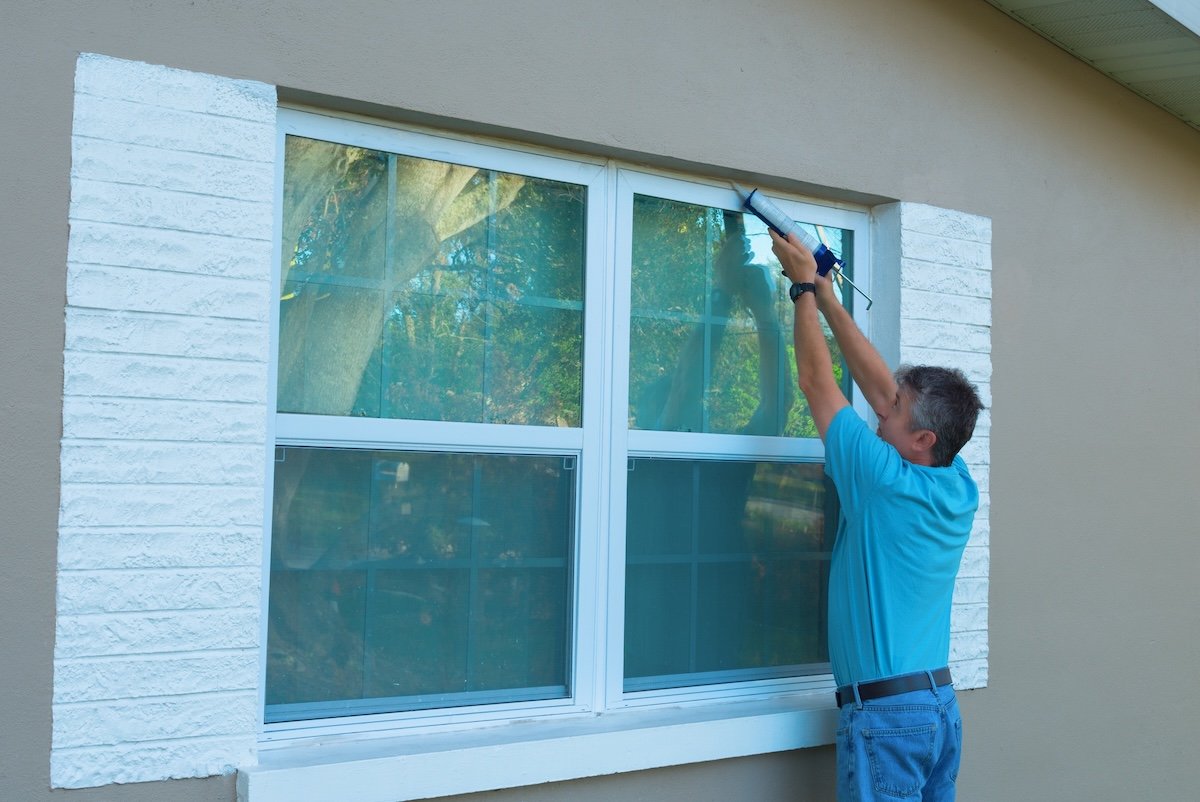 Male homeowner caulking exterior window of a stucco home with a caulk gun.