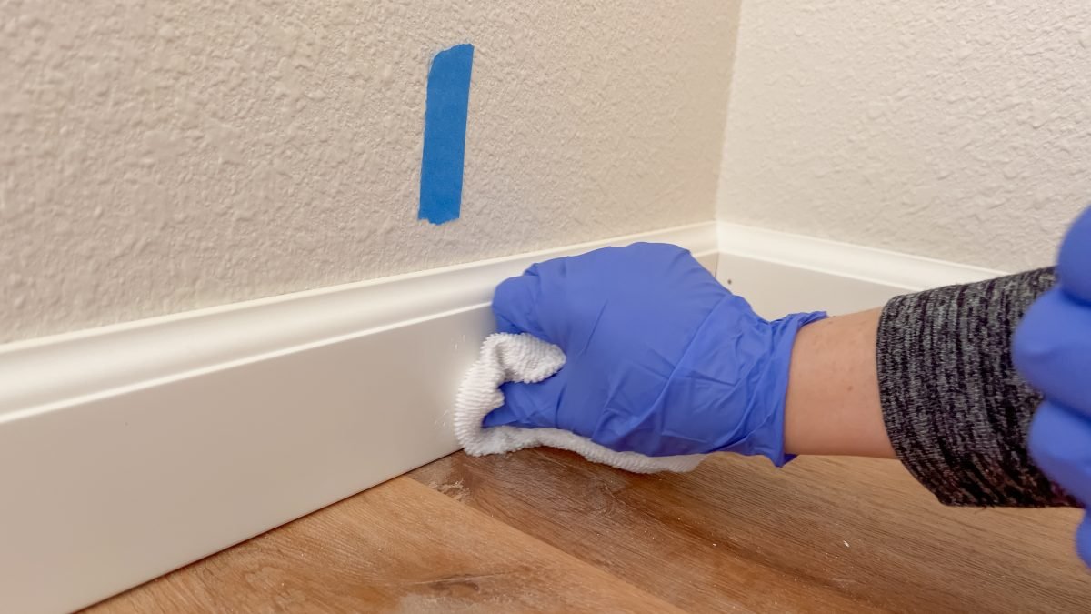 Gloved hand wipes a white baseboard with a cloth, preparing it for painting.