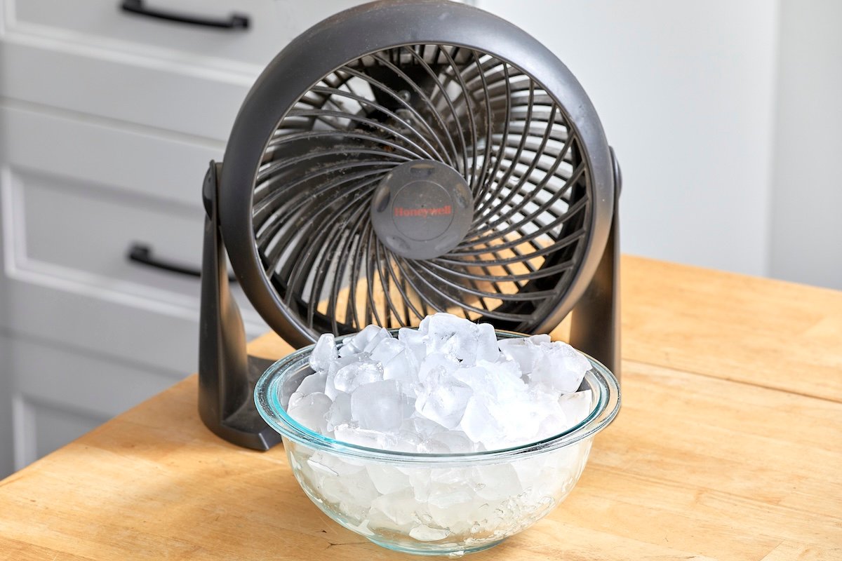 A tabletop fan sits behind a bowl of ice on a table.
