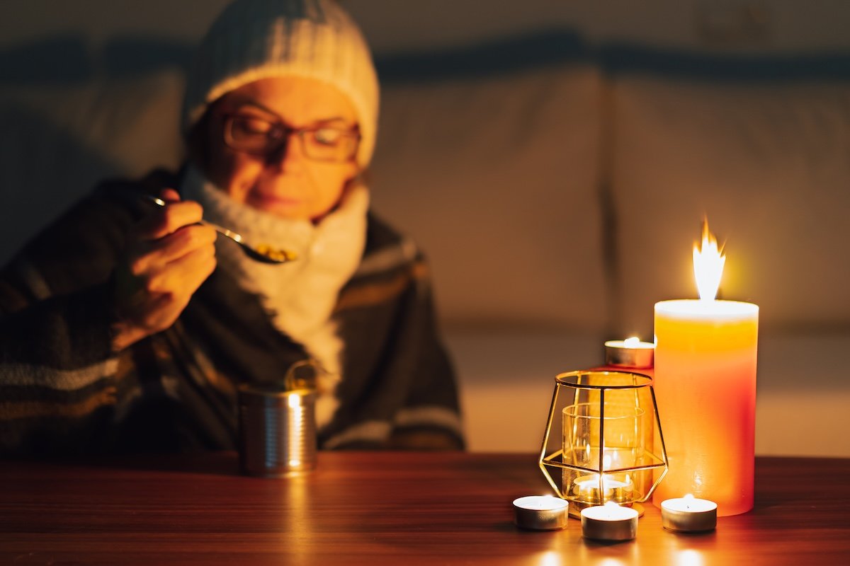 Woman wearing scarf and hat in candlelight, eating emergency food.