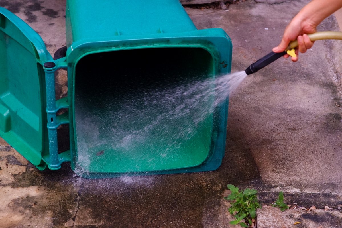 A person is using the water hose to clean the outdoor trash can.