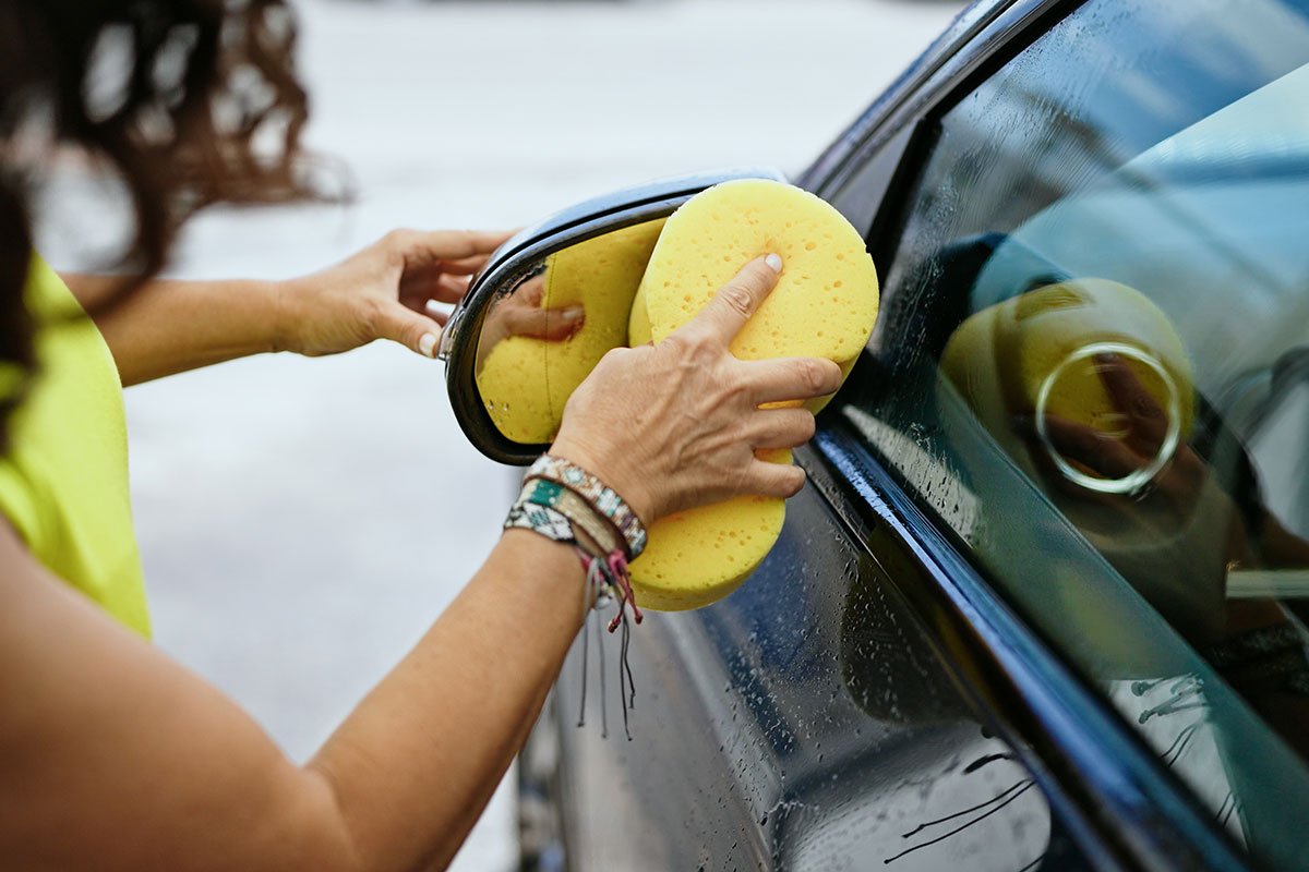 A woman cleaning the side mirror on her car for weekly maintenance.