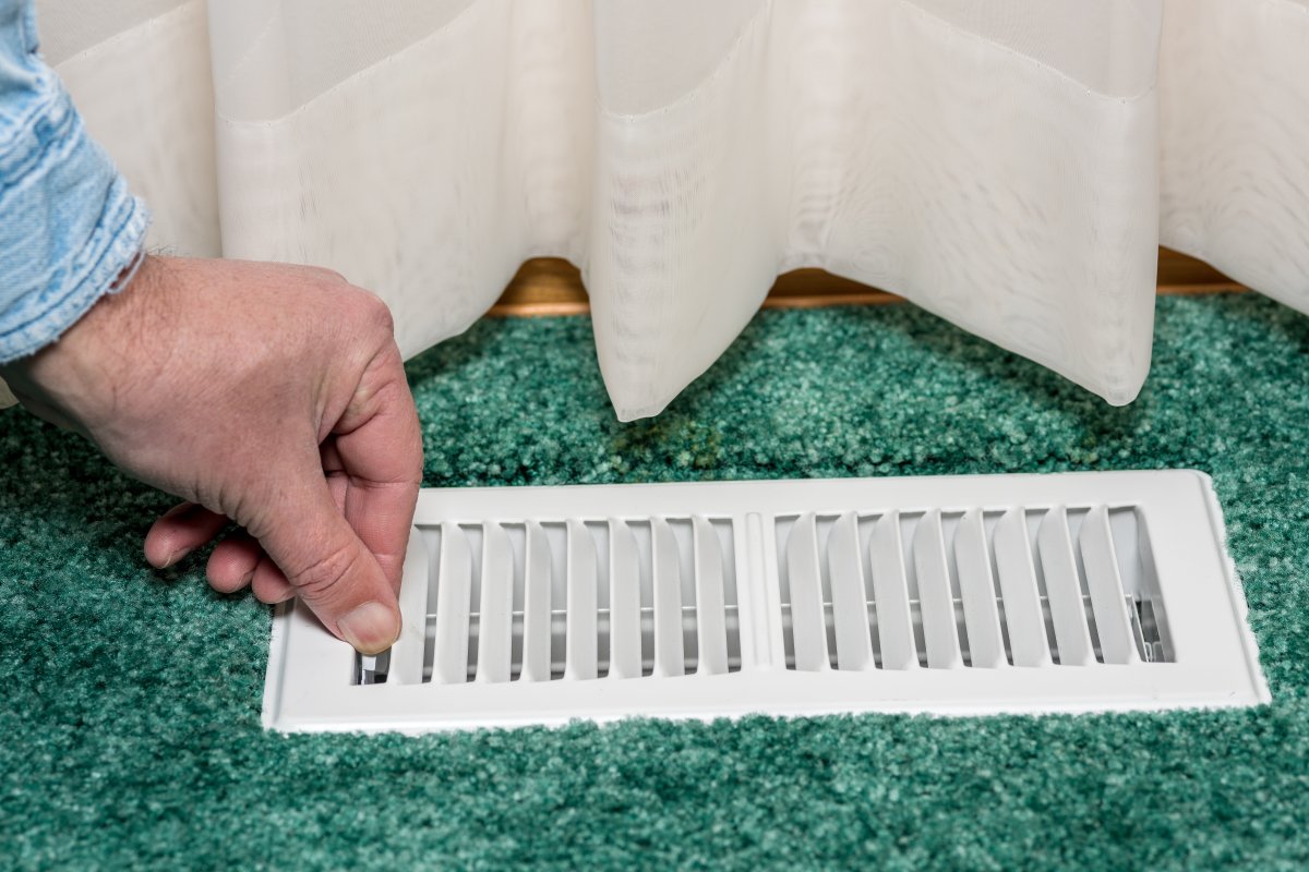 Closeup of person's hand closing louvers on a floor vent, teal carpet and beige drapes in frame.