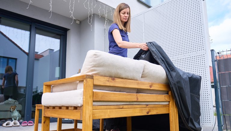 Woman handles black protective cover on wooden patio sofa with cushions outside home.