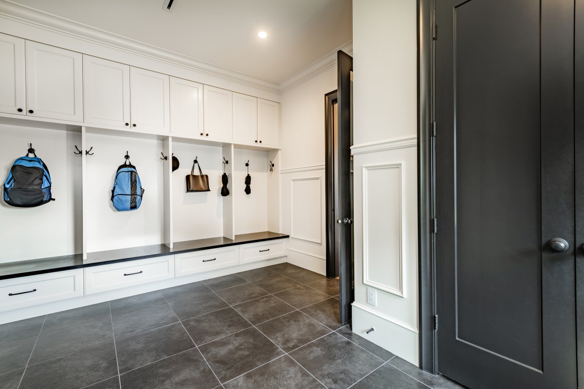 A few backpacks hang in a large, minimalist mudroom with white paneling and dark floors.