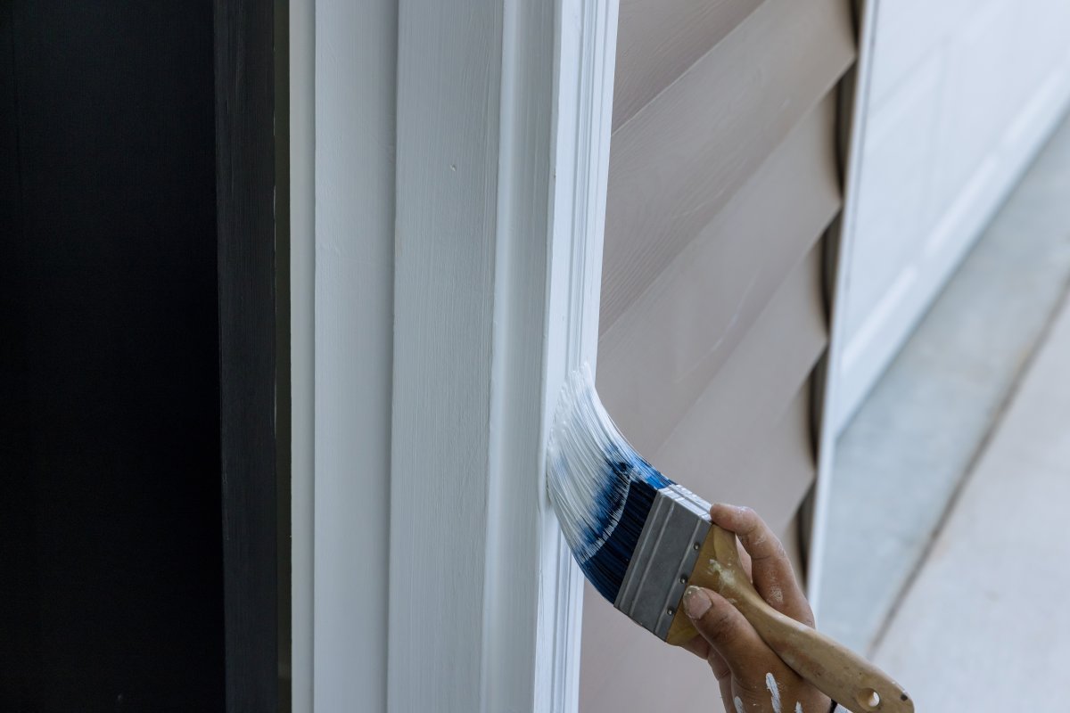 Person uses paint brush to paint white trim around the front door of a house with beige siding.