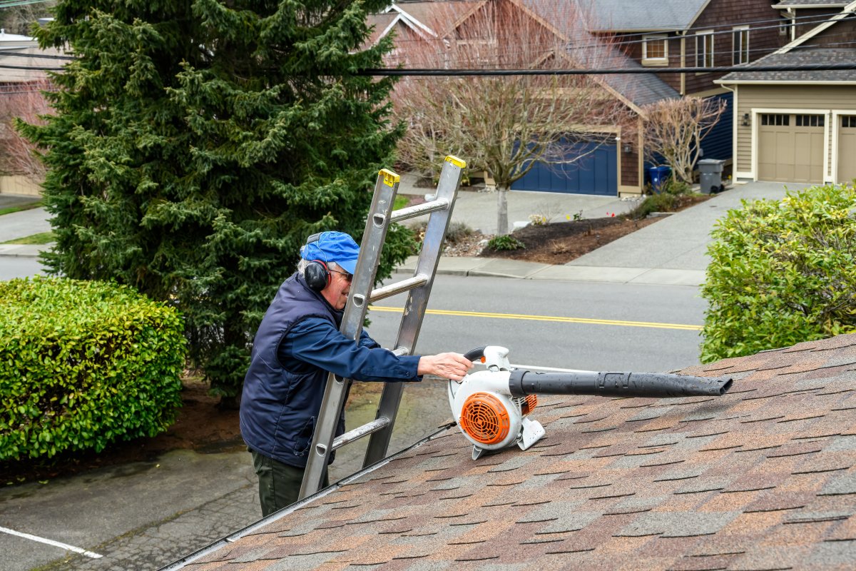 Man climbing an aluminum ladder up to a rooftop with a gas powered leaf blower to clear gutters.
