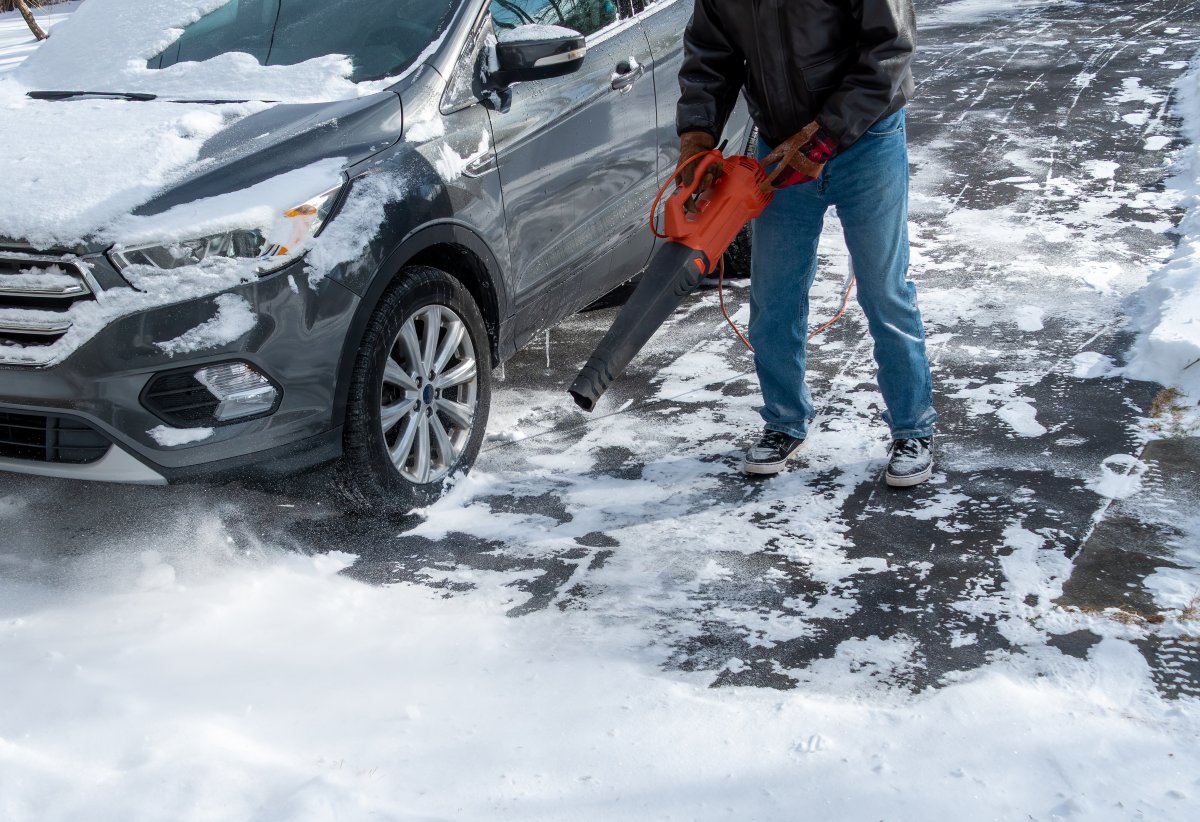 A man using a leaf blower to clear light powdery snow from car and the driveway