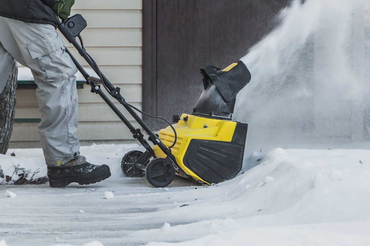 A person uses a yellow and black snow thrower to clear a short walkway. 