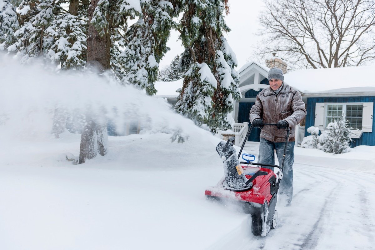 A man wearing snow gear uses a snow blower to clear his snowy driveway.