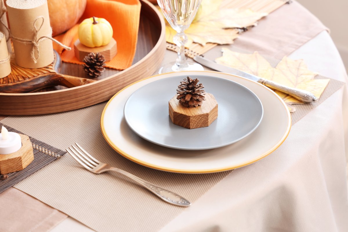 A pinecone sits atop a slice of wood at a Thanksgiving place setting, plate on top of charger and elegant surroundings.